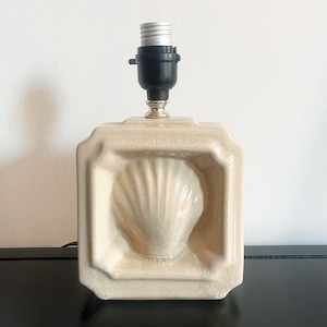 Può includere: Lampada in ceramica color crema con design a conchiglia. La lampada ha una base quadrata con un motivo a conchiglia incorniciato e un portalampada nero. La lampada è su una superficie nera contro un muro bianco.