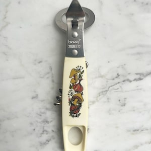 May include: A vintage Bonny Stainless combination bottle opener and corkscrew. The off-white handle features a colorful illustration of mushrooms and onions. The opener has a silver metal finish and a built-in corkscrew.
