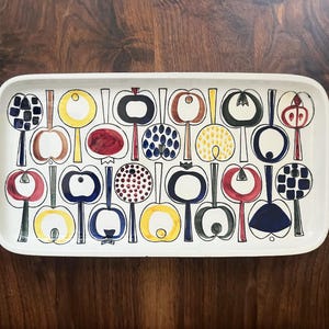 Puede incluir: Bandeja de servicio rectangular blanca con un diseño de manzanas coloridas. Las manzanas son de varios colores, incluyendo rojo, amarillo, azul y negro, con tallos negros. La bandeja está sobre una superficie de madera oscura.