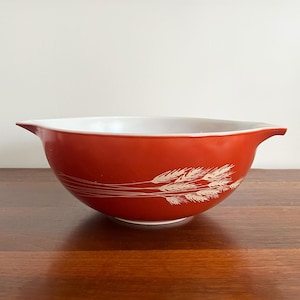 Puede incluir: Un cuenco rojo con interior blanco y diseño de espigas de trigo. El cuenco tiene forma redondeada con borde acampanado y dos asas pequeñas. El diseño de trigo es blanco.