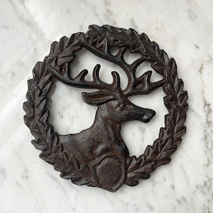Puede incluir: Cabeza de ciervo de hierro fundido dentro de una corona de laurel. El ciervo se representa de perfil, con astas detalladas y un elemento en forma de escudo en el pecho. La corona y el ciervo son de color marrón oscuro.