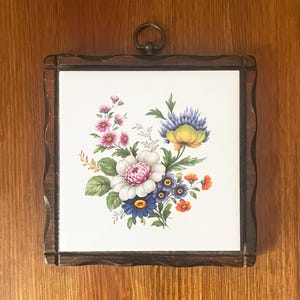 以下が含まれることがあります： ピンク、白、青、オレンジの花と緑の葉が描かれたカラフルな花のイラストが施された正方形のセラミックタイル。ダークブラウンの波状の木製フレームに収められており、上部中央には金属製の吊り下げ用リングが付いています。