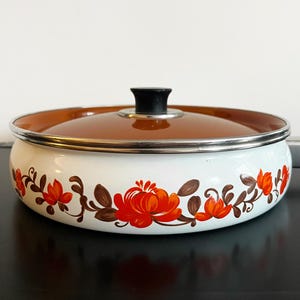May include: A white enamel cooking pot with a brown lid and a black handle. The pot is decorated with a floral pattern in shades of red and brown. The pot has a silver rim.