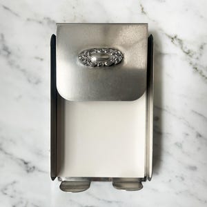 Könnte beinhalten: Ein Tissuebox-Halter aus gebürstetem Silber mit einem verzierten ovalen Schmuck. Der Halter hat einen Klappdeckel und eine rechteckige Öffnung zur Ausgabe von Taschentüchern. Das Design ist schlicht und elegant.