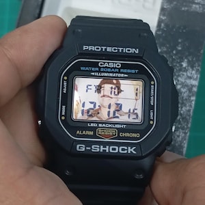 May include: A black Casio G-Shock digital watch with a square face, featuring the text PROTECTION, WATER 20BAR RESIST, ILLUMINATOR, LED BACKLIGHT, ALARM, SHOCK RESIST, and CHRONO. The digital display shows the time 12:12:15 and date FR 10-1.