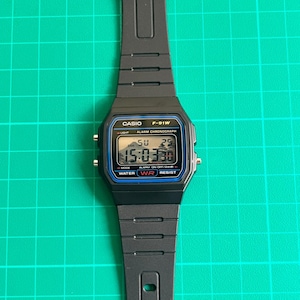 K&ouml;nnte beinhalten: Schwarze digitale Casio F-91W-Uhr mit schwarzem Harzarmband. Das Zifferblatt zeigt die Uhrzeit, den Tag und das Datum an. Auf der Uhr steht "CASIO F-91W" und "WATER RESIST". Das Armband ist ca. 24 cm lang.