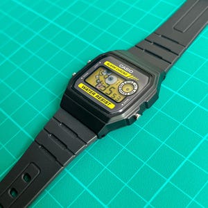 May include: Black Casio digital watch with a yellow display featuring a cartoon character. The watch has a black band and a water-resistant label. The watch face displays the time and other functions. The watch is on a green surface.