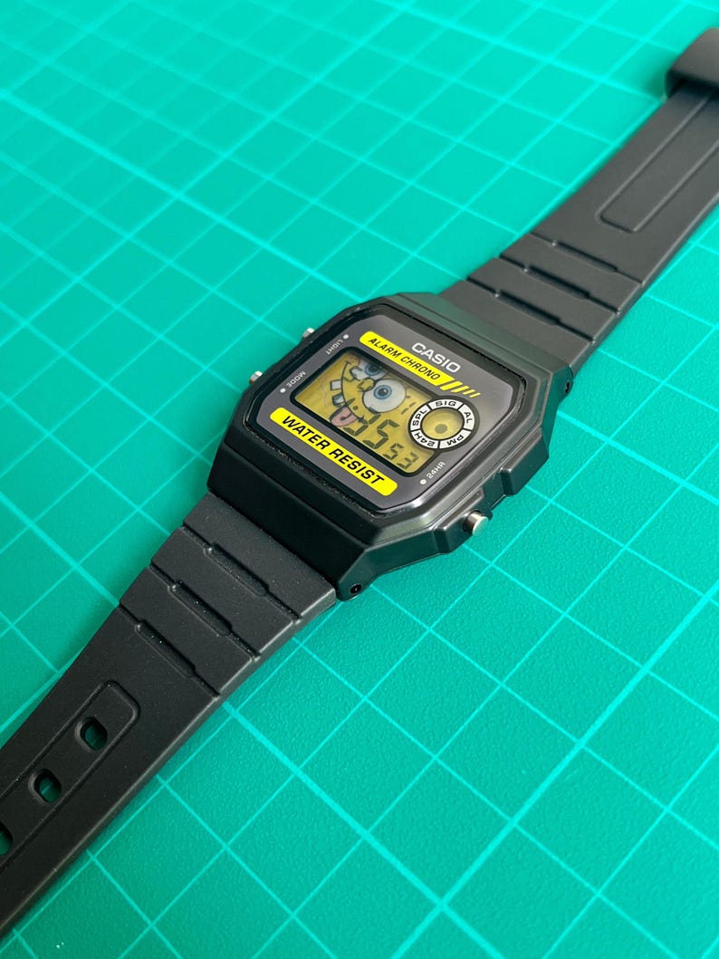 May include: Black Casio digital watch with a yellow display featuring a cartoon character. The watch has a black band and a water-resistant label. The watch face displays the time and other functions. The watch is on a green surface.