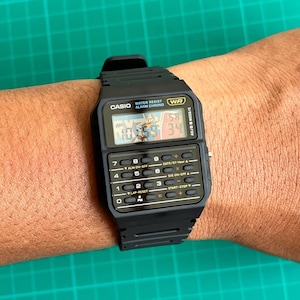 May include: Black Casio digital watch with a rectangular face and multiple buttons. The watch displays the time and has the text "WATER RESIST ALARM CHRONO" and "WR". The watch is on a wrist.
