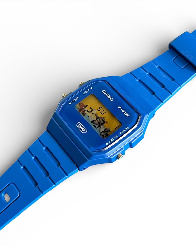 May include: A vibrant blue Casio F-91W digital watch. The watch features a rectangular face with a digital display showing the time. The watchband and case are a matching bright blue. The watch has buttons for mode, light, and alarm functions.