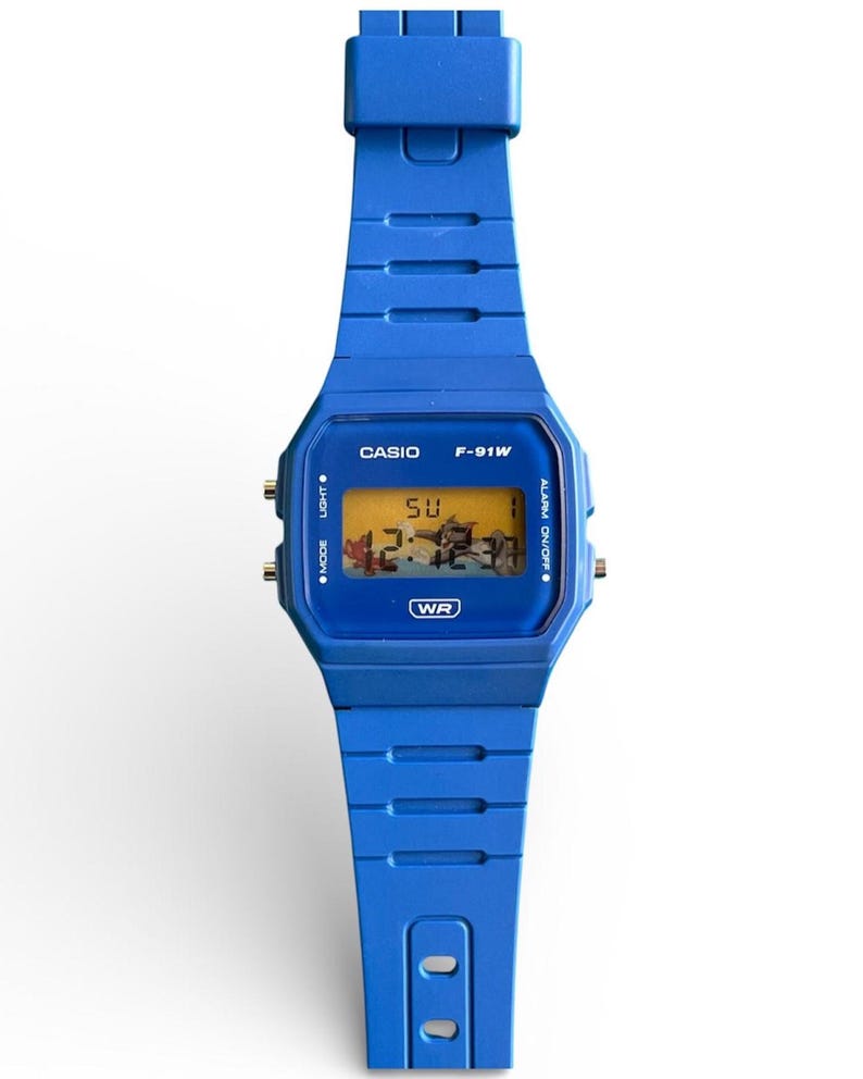 May include: A blue Casio F-91W digital watch. The watch features a blue resin strap and case, with a digital display showing the time and day. The watch face displays the text "CASIO" and "F-91W". The watch is water resistant.