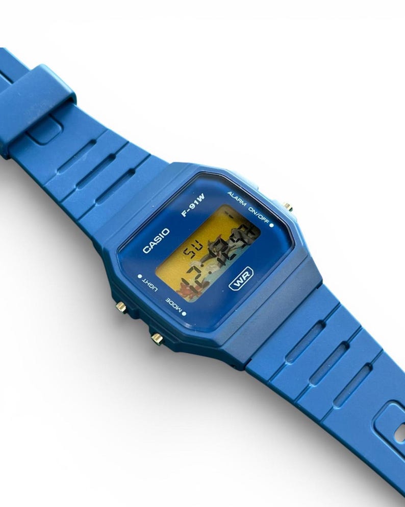 May include: A blue Casio F-91W digital watch with a matching blue strap. The watch face displays the time and day. The watch has the text "CASIO", "F-91W", "WR", "LIGHT", "MODE", and "ALARM ON/OFF".