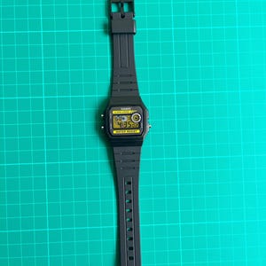 May include: Black digital watch with a rectangular face and a black band. The watch face displays the time in yellow digits. The watch has the word "CASIO" printed on the face and the words "WATER RESIST" below the display.