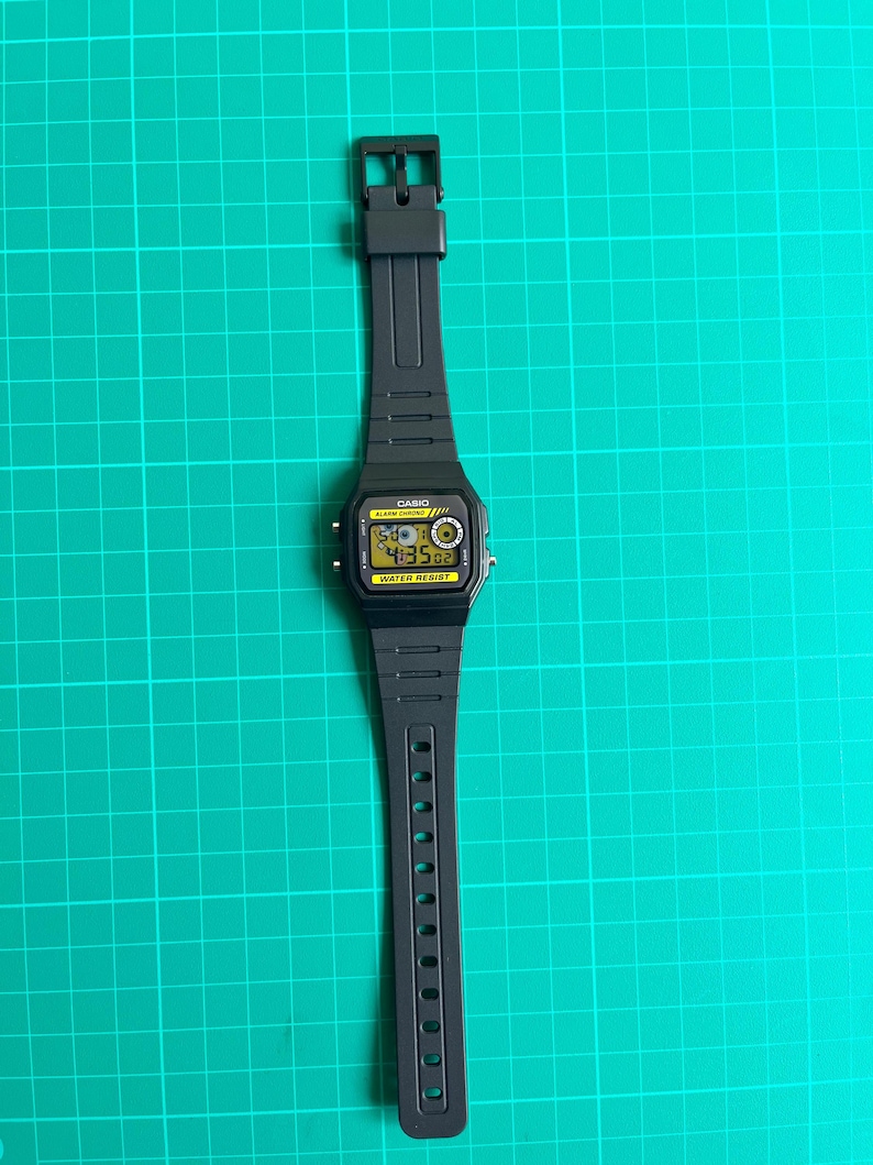 May include: Black digital watch with a rectangular face and a black band. The watch face displays the time in yellow digits. The watch has the word "CASIO" printed on the face and the words "WATER RESIST" below the display.
