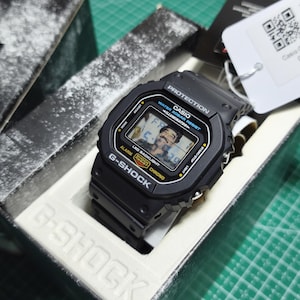 May include: A black Casio G-Shock digital watch with a square face, featuring the text PROTECTION, WATER 20BAR RESIST, ILLUMINATOR, LED BACKLIGHT, SHOCK RESIST, and CHRONO. The watch display shows a digital image of a person with a mustache.