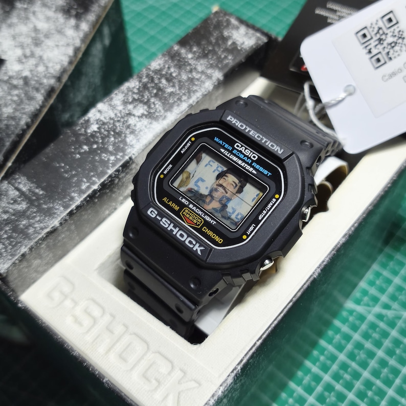 May include: A black Casio G-Shock digital watch with a square face, featuring the text PROTECTION, WATER 20BAR RESIST, ILLUMINATOR, LED BACKLIGHT, SHOCK RESIST, and CHRONO. The watch display shows a digital image of a person with a mustache.