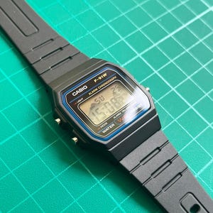 K&ouml;nnte beinhalten: Eine schwarze Casio F-91W Digitaluhr mit rechteckigem Zifferblatt und schwarzem Armband. Die Uhr zeigt Uhrzeit, Datum und andere Funktionen an. Die Uhr hat einen blauen und silbernen Rand um den Bildschirm. Die Uhr ist wasserdicht.