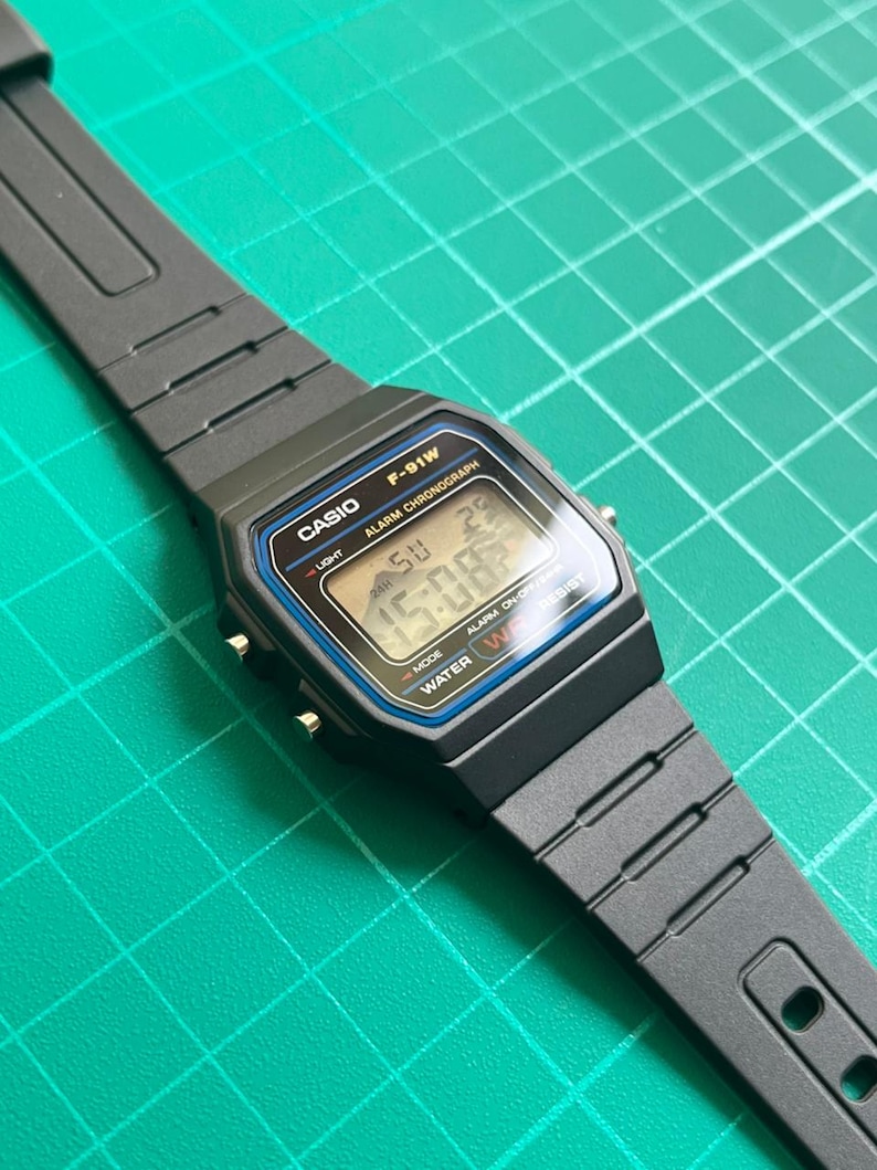 K&ouml;nnte beinhalten: Eine schwarze Casio F-91W Digitaluhr mit rechteckigem Zifferblatt und schwarzem Armband. Die Uhr zeigt Uhrzeit, Datum und andere Funktionen an. Die Uhr hat einen blauen und silbernen Rand um den Bildschirm. Die Uhr ist wasserdicht.