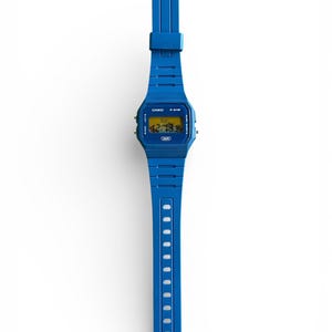 May include: A blue digital wristwatch with a matching blue band. The watch face displays the time and day in yellow digits. The watch band has a series of holes for adjustment. The watch is a Casio F-91W model.