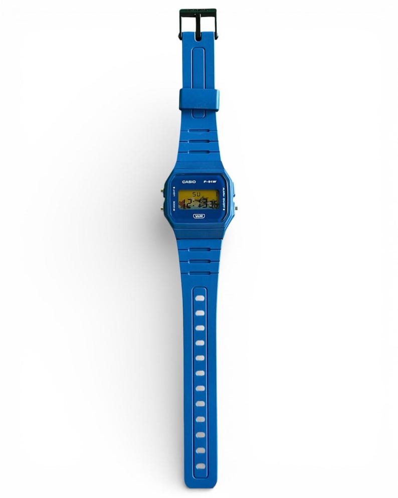 May include: A blue digital wristwatch with a matching blue band. The watch face displays the time and day in yellow digits. The watch band has a series of holes for adjustment. The watch is a Casio F-91W model.