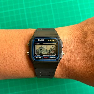 K&ouml;nnte beinhalten: Eine schwarze Casio F-91W Digitaluhr mit rechteckigem Zifferblatt und schwarzem Harzarmband. Die Uhr zeigt die Uhrzeit, den Tag und das Datum an. Das Zifferblatt hat blaue und silberne Akzente und der Text "CASIO F-91W" ist sichtbar.