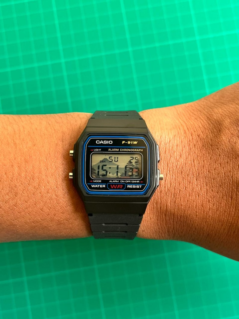 K&ouml;nnte beinhalten: Eine schwarze Casio F-91W Digitaluhr mit rechteckigem Zifferblatt und schwarzem Harzarmband. Die Uhr zeigt die Uhrzeit, den Tag und das Datum an. Das Zifferblatt hat blaue und silberne Akzente und der Text "CASIO F-91W" ist sichtbar.