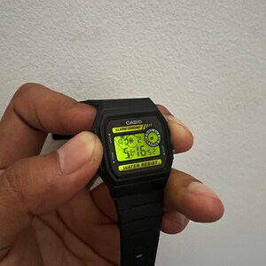 May include: Black Casio digital watch with a rectangular face and black strap. The watch face displays the time and date in a green digital format. The watch has the text "CASIO" and "WATER RESIST" on the face.