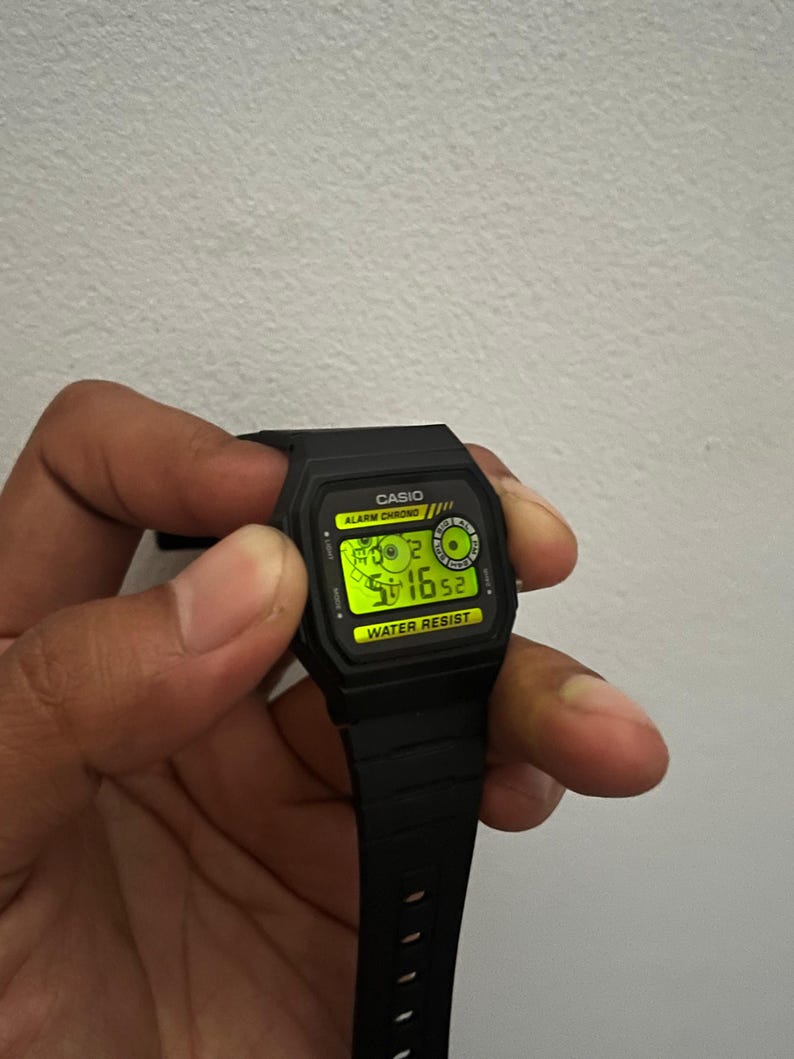 May include: Black Casio digital watch with a rectangular face and black strap. The watch face displays the time and date in a green digital format. The watch has the text "CASIO" and "WATER RESIST" on the face.
