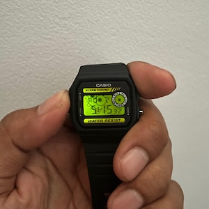 May include: A black Casio digital watch with a rectangular face and a black strap. The watch face displays the time in green digits. The watch has the words "CASIO", "ALARM CHRONO", and "WATER RESIST" printed on the face.