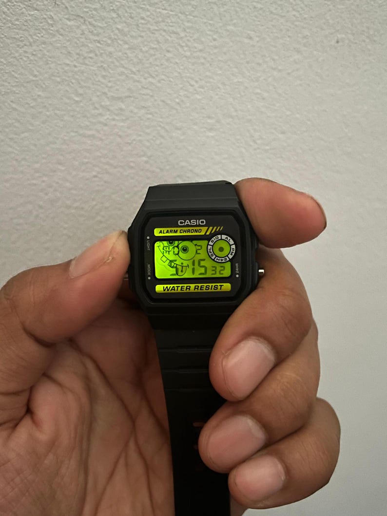 May include: A black Casio digital watch with a rectangular face and a black strap. The watch face displays the time in green digits. The watch has the words "CASIO", "ALARM CHRONO", and "WATER RESIST" printed on the face.