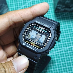 May include: A black Casio G-Shock digital watch with a rectangular face. The screen displays the text PROTECTION, WATER 20BAR RESIST, ILLUMINATOR, ALARM, SHOCK RESIST, and CHRONO, with a small portrait of a man visible on the digital display.