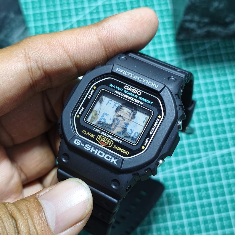 May include: A black Casio G-Shock digital watch with a rectangular face. The screen displays the text PROTECTION, WATER 20BAR RESIST, ILLUMINATOR, ALARM, SHOCK RESIST, and CHRONO, with a small portrait of a man visible on the digital display.
