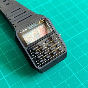May include: A black Casio digital watch with a rectangular face and integrated calculator buttons. The watch displays the time and has a black rubber strap. The watch face includes the text "CASIO WATER RESIST ALARM CHRONO".