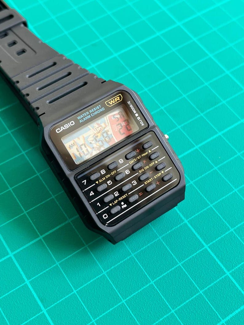 May include: A black Casio digital watch with a rectangular face and integrated calculator buttons. The watch displays the time and has a black rubber strap. The watch face includes the text "CASIO WATER RESIST ALARM CHRONO".