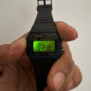 K&ouml;nnte beinhalten: Schwarze Casio F-91W Digitaluhr mit rechteckigem Zifferblatt, das die Uhrzeit, den Tag und das Datum in Gr&uuml;n anzeigt. Die Uhr hat ein schwarzes Harzarmband und wird von einer Hand gehalten. Das Zifferblatt tr&auml;gt die Aufschrift "CASIO F-91W" und "WATER RESIST".