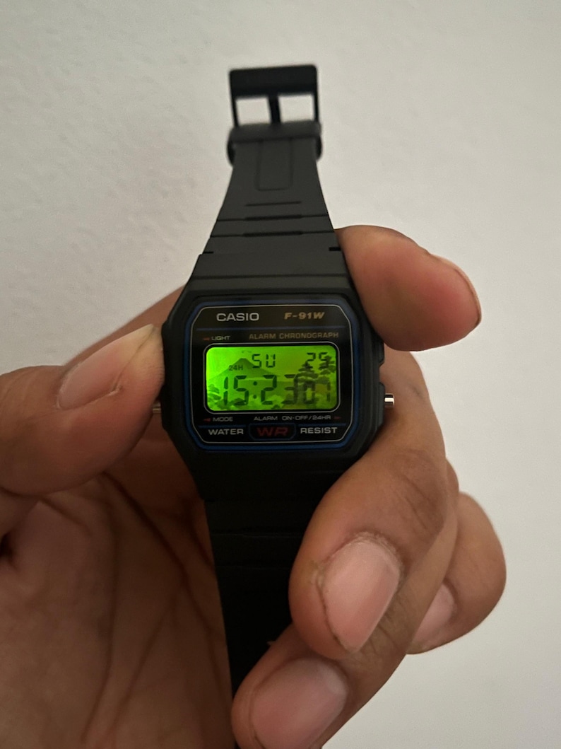 K&ouml;nnte beinhalten: Schwarze Casio F-91W Digitaluhr mit rechteckigem Zifferblatt, das die Uhrzeit, den Tag und das Datum in Gr&uuml;n anzeigt. Die Uhr hat ein schwarzes Harzarmband und wird von einer Hand gehalten. Das Zifferblatt tr&auml;gt die Aufschrift "CASIO F-91W" und "WATER RESIST".