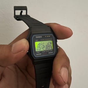 K&ouml;nnte beinhalten: Eine schwarze Casio F-91W Digitaluhr mit schwarzem Armband. Das Zifferblatt zeigt die Uhrzeit, den Tag und das Datum in gr&uuml;nen Ziffern an. Auf der Uhr sind der Name Casio und das Modell F-91W aufgedruckt. Die Uhr wird von einer Hand gehalten.