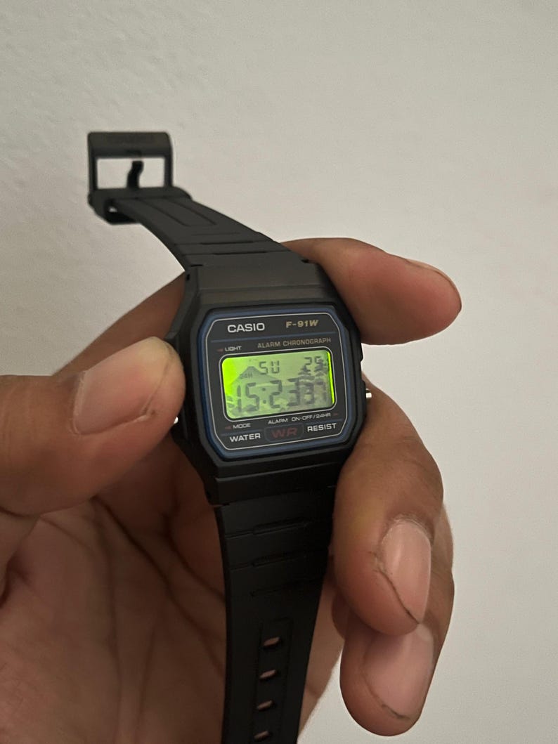 K&ouml;nnte beinhalten: Eine schwarze Casio F-91W Digitaluhr mit schwarzem Armband. Das Zifferblatt zeigt die Uhrzeit, den Tag und das Datum in gr&uuml;nen Ziffern an. Auf der Uhr sind der Name Casio und das Modell F-91W aufgedruckt. Die Uhr wird von einer Hand gehalten.