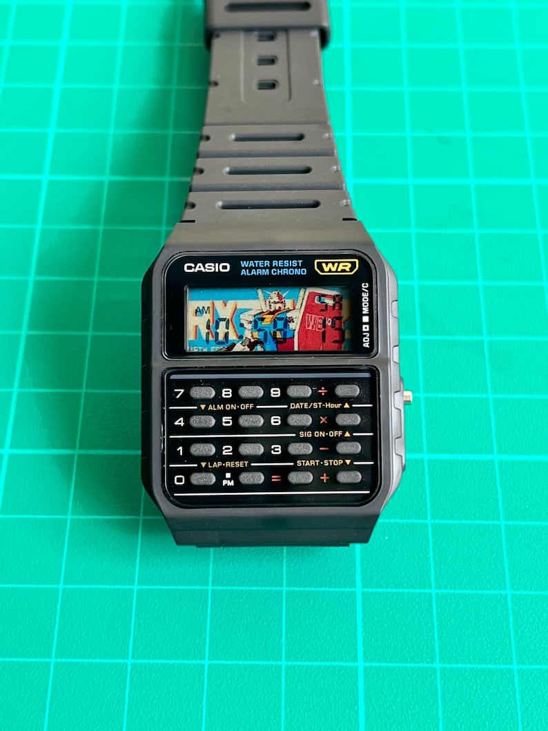 May include: A dark gray Casio digital watch with a calculator keypad and a small screen displaying a colorful graphic. The watch band is also dark gray. The watch face has the text "CASIO WATER RESIST ALARM CHRONO WR".