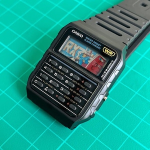 May include: A black Casio digital watch with a calculator keypad and a gray strap. The watch face displays a colorful graphic and the text "RX". Features include water resistance and an alarm function.