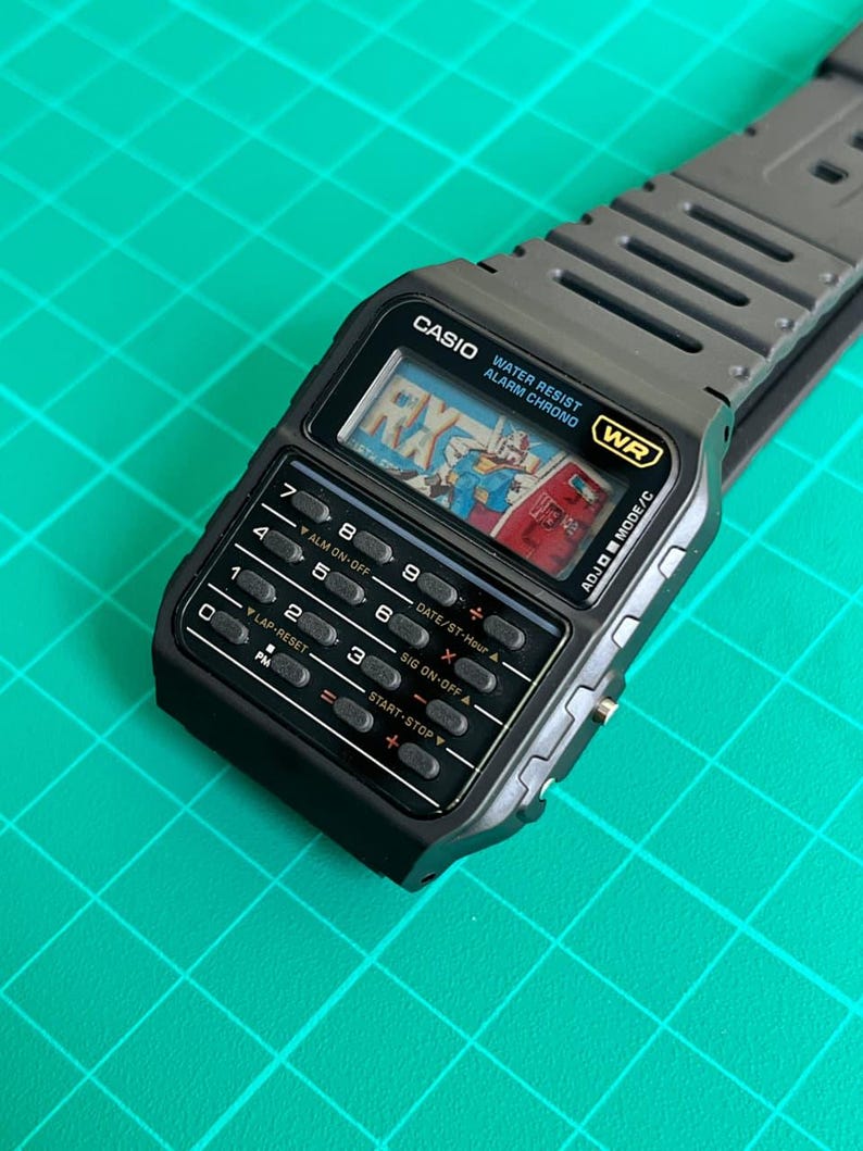 May include: A black Casio digital watch with a calculator keypad and a gray strap. The watch face displays a colorful graphic and the text "RX". Features include water resistance and an alarm function.