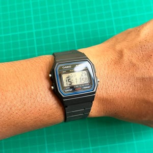 K&ouml;nnte beinhalten: Schwarze Digitaluhr mit rechteckigem Zifferblatt und schwarzem Armband. Das Zifferblatt zeigt die Uhrzeit, den Tag und das Datum an. Die Uhr hat den Markennamen "CASIO" oben und den Text "WATER WR RESIST" unten.