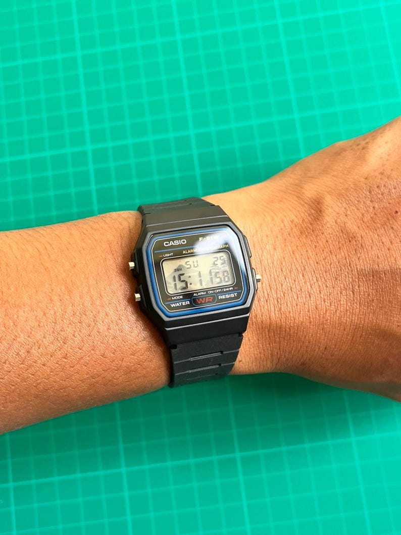 K&ouml;nnte beinhalten: Schwarze Digitaluhr mit rechteckigem Zifferblatt und schwarzem Armband. Das Zifferblatt zeigt die Uhrzeit, den Tag und das Datum an. Die Uhr hat den Markennamen "CASIO" oben und den Text "WATER WR RESIST" unten.
