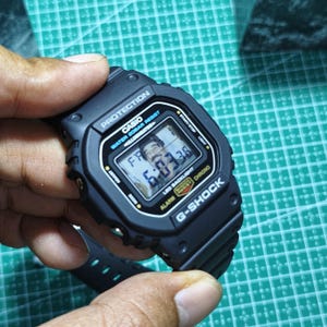 May include: A black Casio G-Shock digital watch with a rectangular face, featuring the text PROTECTION, WATER 20BAR RESIST, ILLUMINATOR, LED BACKLIGHT, and G-SHOCK. The digital display shows the time 6:03:38 and the day FR.