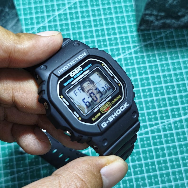 May include: A black Casio G-Shock digital watch with a rectangular face, featuring the text PROTECTION, WATER 20BAR RESIST, ILLUMINATOR, LED BACKLIGHT, and G-SHOCK. The digital display shows the time 6:03:38 and the day FR.