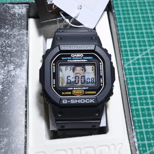 May include: A black Casio G-Shock digital watch with a square face, featuring the text PROTECTION, CASIO, WATER 20BAR RESIST, ILLUMINATOR, LED BACKLIGHT, ALARM, SHOCK RESIST, CHRONO, and G-SHOCK. The screen displays the time 6:00:08 and the date FR 1.