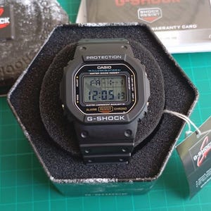 May include: Black G-Shock digital watch with a rectangular face, presented in its original hexagonal box. The watch face displays the time, day, and date. The watch has multiple buttons and the text "G-Shock" is visible.
