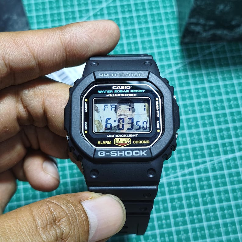 May include: A black Casio G-Shock digital watch with a square face. The screen displays the time 6:03:50, the date FR 1, and a small portrait of a man. Text on the bezel includes PROTECTION, WATER 20BAR RESIST, ILLUMINATOR, and G-SHOCK.