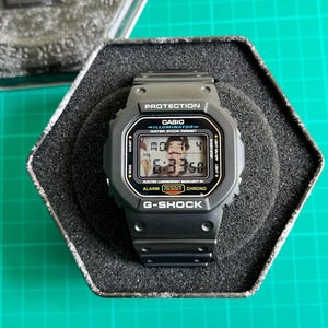 Könnte beinhalten: Eine schwarze G-Shock Digitaluhr mit rechteckigem Zifferblatt, präsentiert in der Originalverpackung. Das Zifferblatt zeigt die Uhrzeit und das Datum an. Die Uhr trägt den Text "CASIO ILLUMINATOR WATER 200M RESIST" und "G-SHOCK".