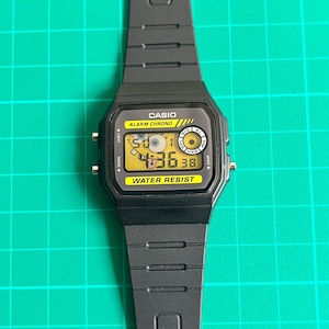 May include: Black Casio digital watch with a rectangular face and a black band. The watch face displays the time and has the words "CASIO", "ALARM CHRONO", and "WATER RESIST" printed on it. The watch is on a green surface.